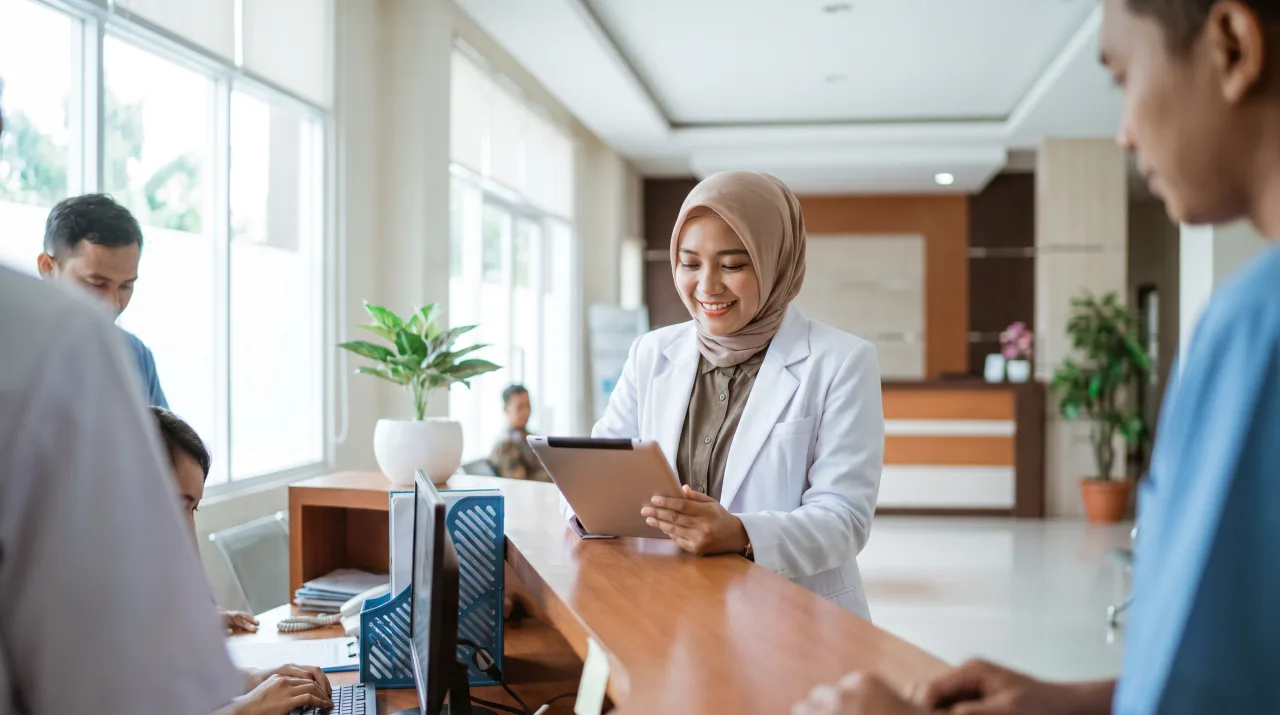 Dokter berjas putih meninjau data pasien lewat tablet di nurse station rumah sakit.